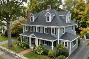 A blue-gray two-story home with a new dark gray shingle roof, multiple dormers, white trim, and mature trees surrounding the property.